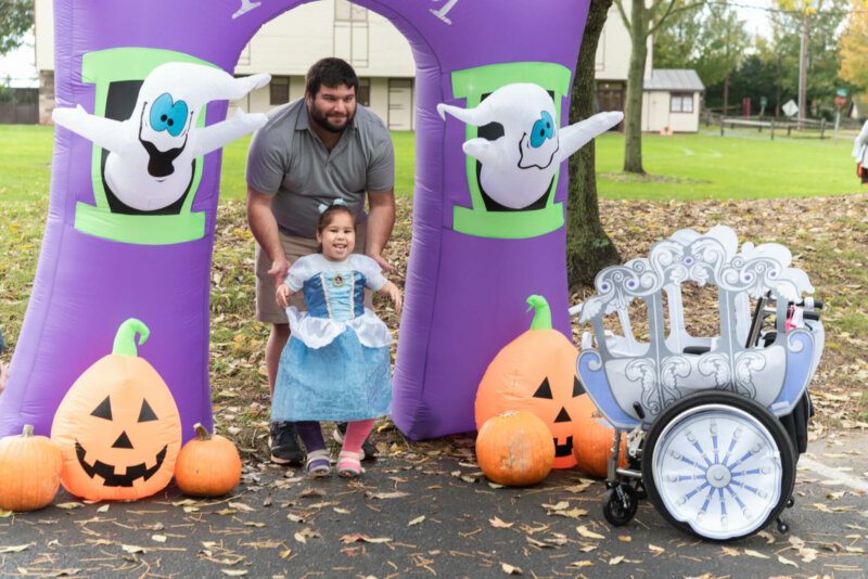 A little girl wearing leg braces dressed as Cinderella poses with her father under an inflatable purple Halloween-themed arch featuring ghosts and jack-o-lanterns. In the right foreground is her wheelchair decorated as Cinderella's Carriage.